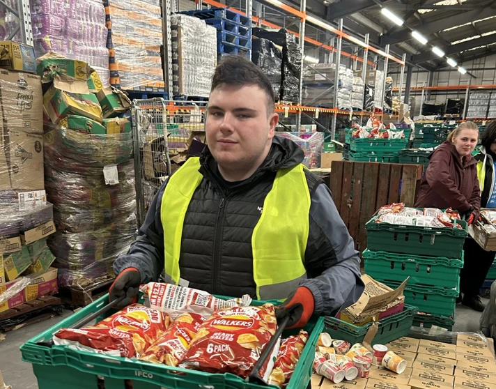 Young man wearing a high-visibility vest holding a crate of packaged crisps while working in a warehouse environment. Young man wearing a high-visibility vest holding a crate of packaged crisps while working in a warehouse environment.